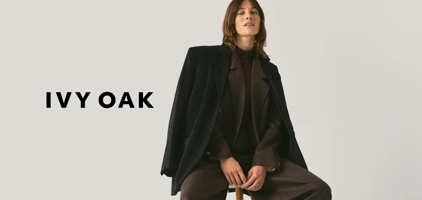 Donna seduta su uno sgabello di legno che indossa pantaloni scuri a gamba larga, top abbinato e giacca nera testurizzata drappeggiata sulle spalle su sfondo semplice.