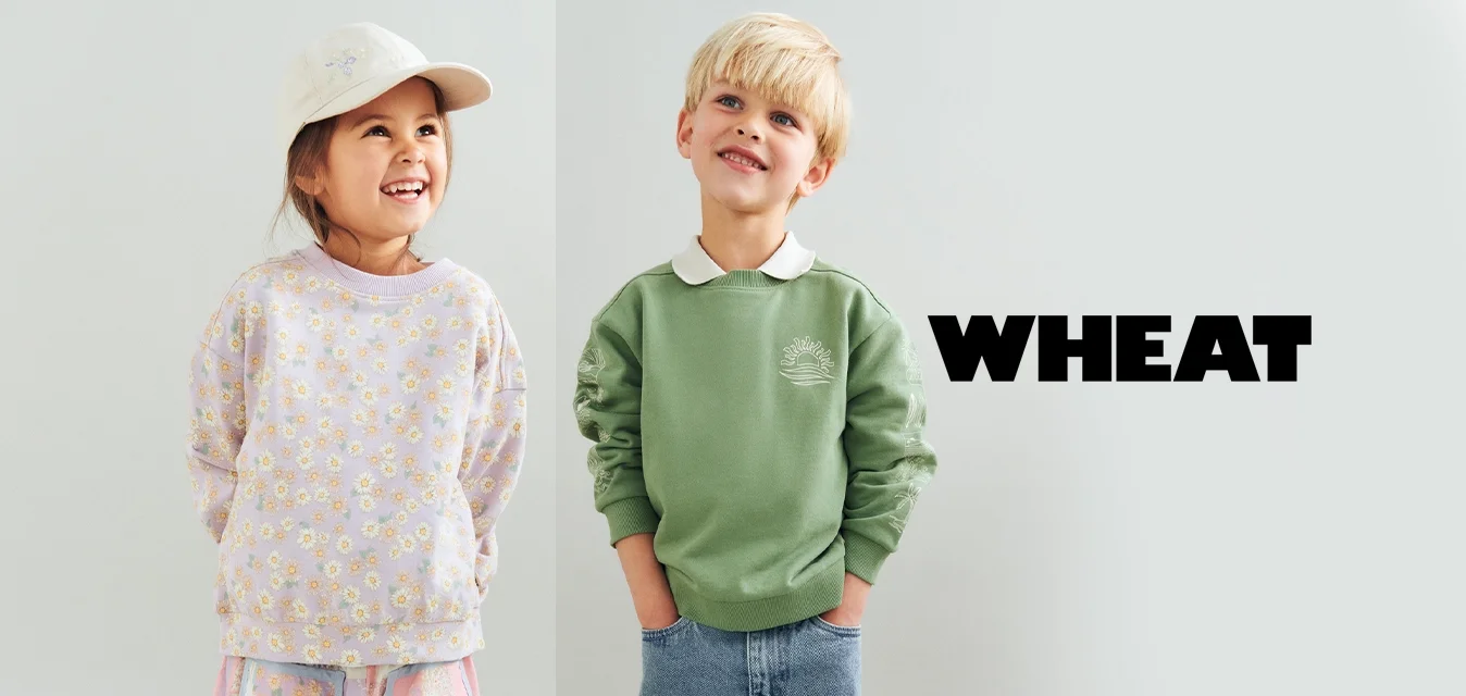 Due bambini sorridenti stanno uno accanto all'altro; la bambina indossa una felpa floreale e un berretto, il bambino indossa una felpa verde e dei jeans.