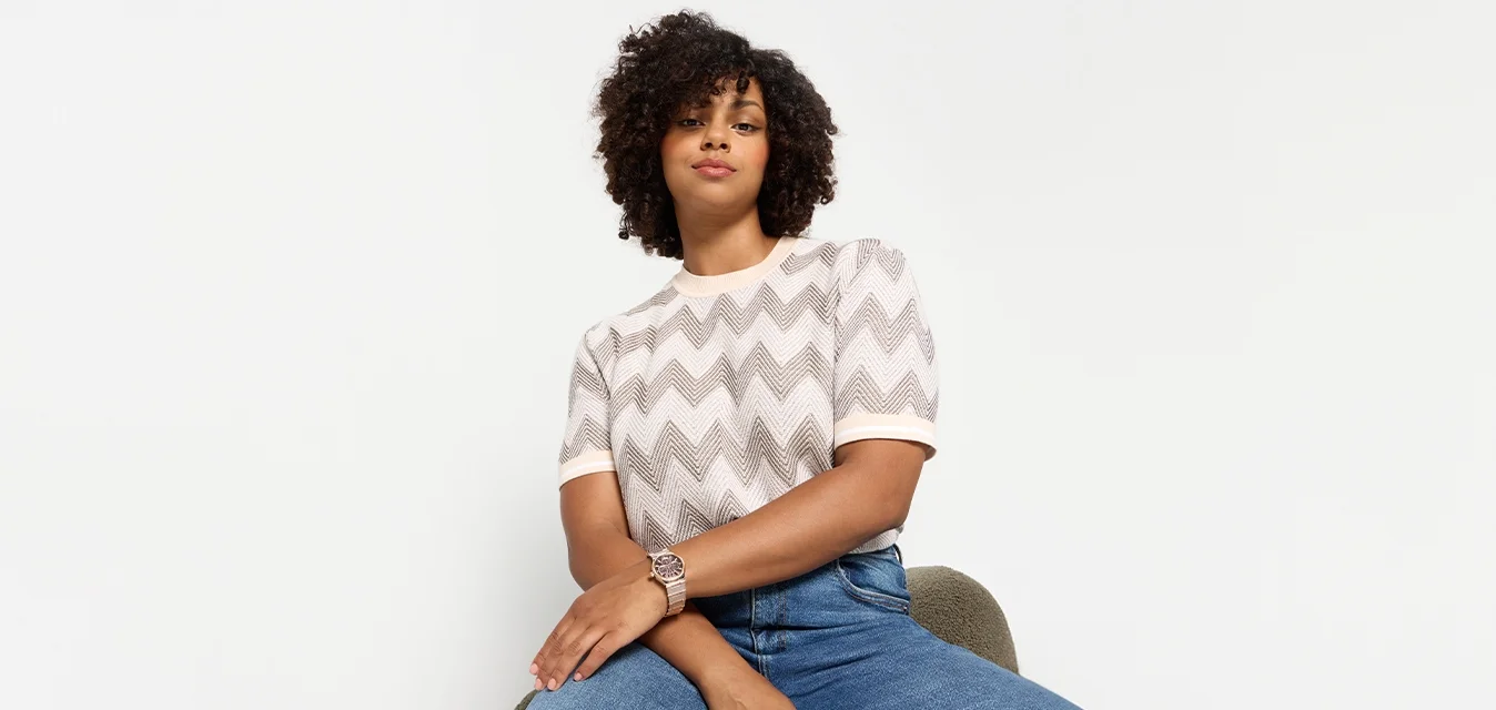 Top a maglia con motivo a zigzag grigio e crema, con maniche corte, abbinato a jeans blu attillati. Seduta su una sedia verde tappezzata.