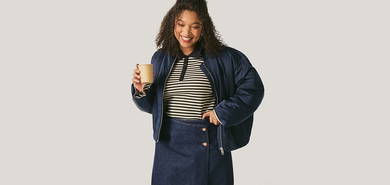 Donna sorridente con giacca blu navy, top a righe e gonna, tiene in mano una tazza di caffè, in piedi davanti a uno sfondo chiaro e semplice.