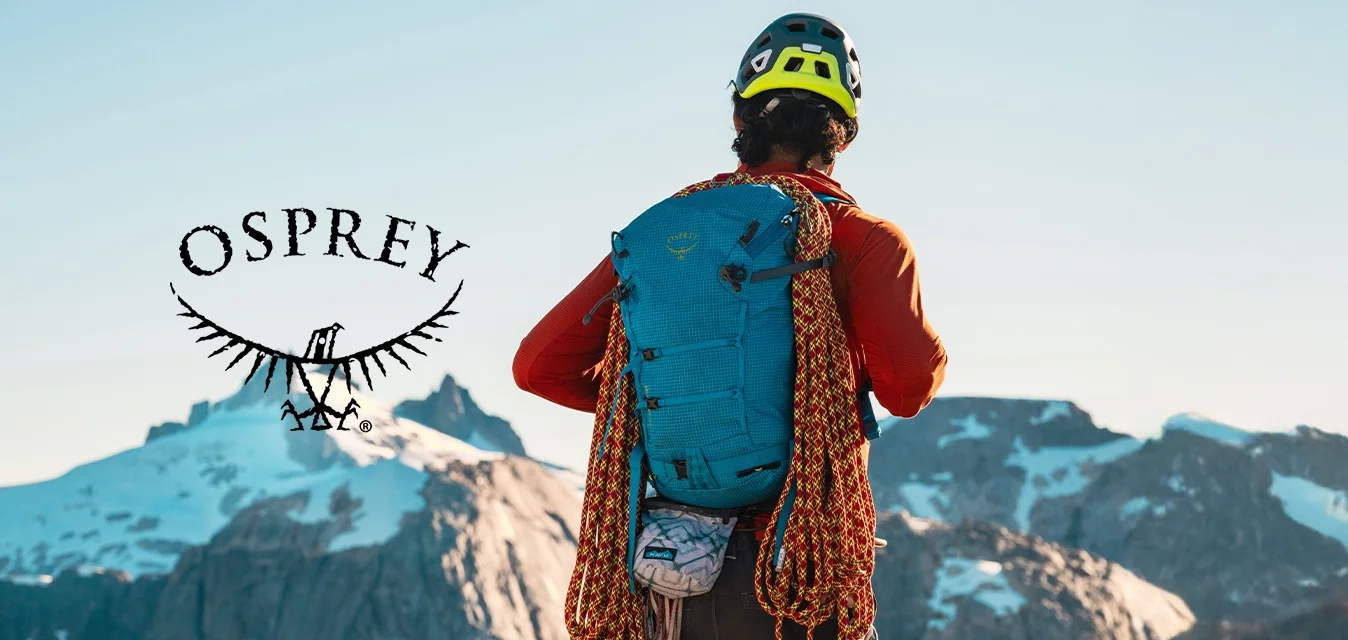 Zaino blu con superficie tessurizzata, corde da arrampicata arancioni attaccate, indossato su una maglietta a maniche lunghe arancione, contro uno sfondo montano.