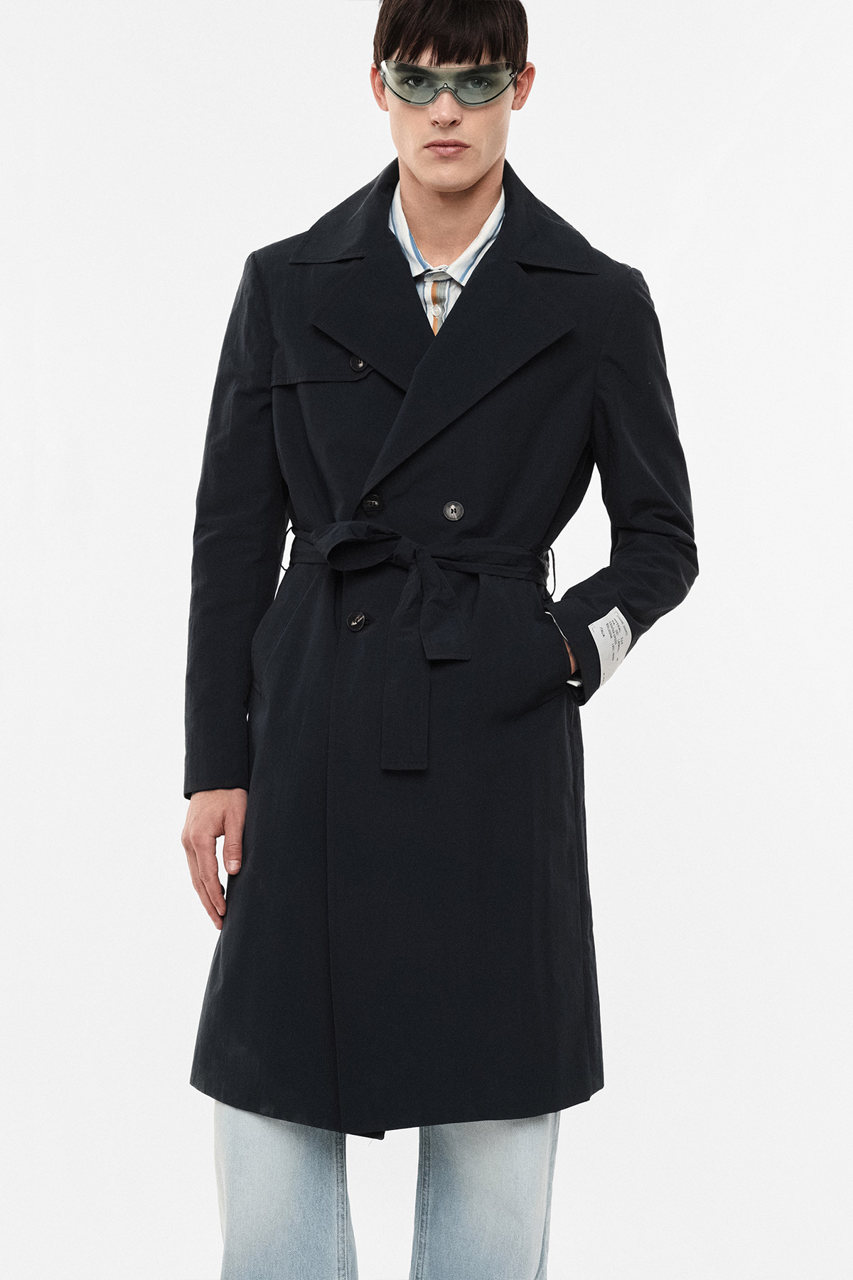 Male model wearing a black belted trench coat over a striped shirt, light-wash jeans and green-tinted sunglasses on a white background