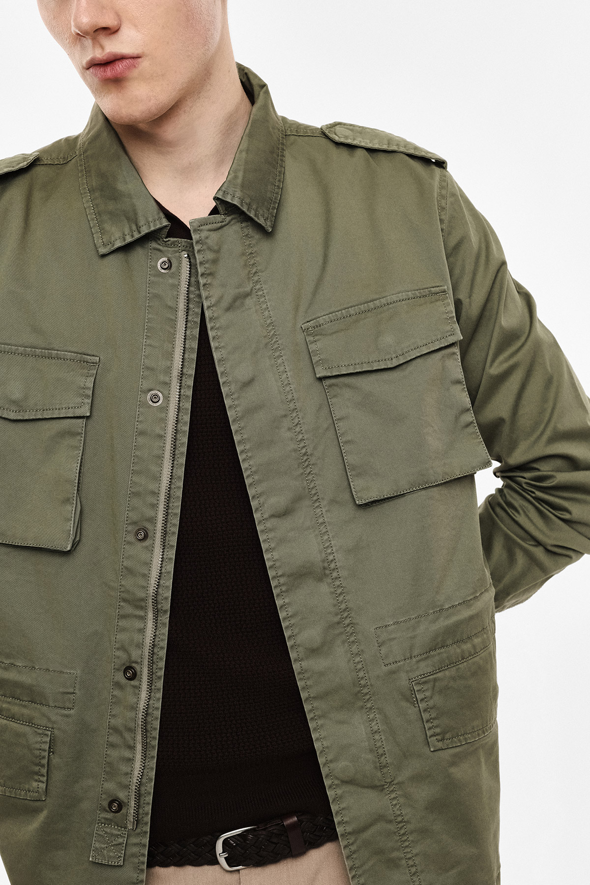 Men's khaki utility jacket with flap pockets layered over a black knit sweater, braided belt visible