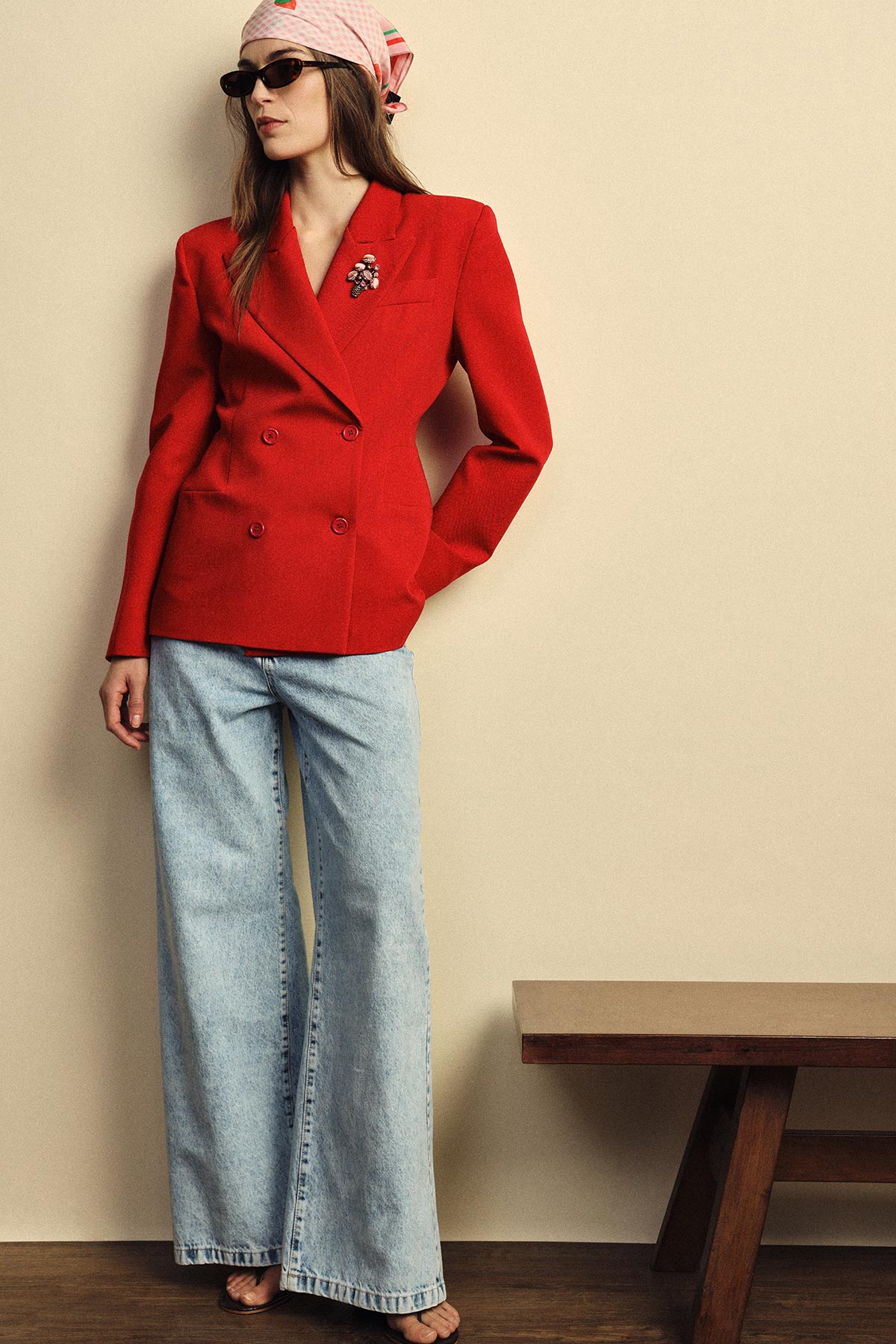 Person in red double-breasted blazer with brooch, pink headscarf, sunglasses and light‑wash wide‑leg jeans leaning by a beige wall