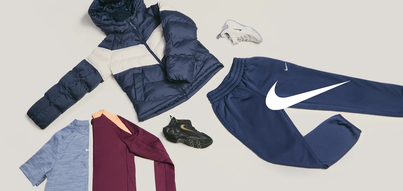 Giacca imbottita blu scuro e beige, camicie blu e bordeaux, pantaloni della tuta blu scuro, scarpe da ginnastica nere e bianche disposti su sfondo chiaro.