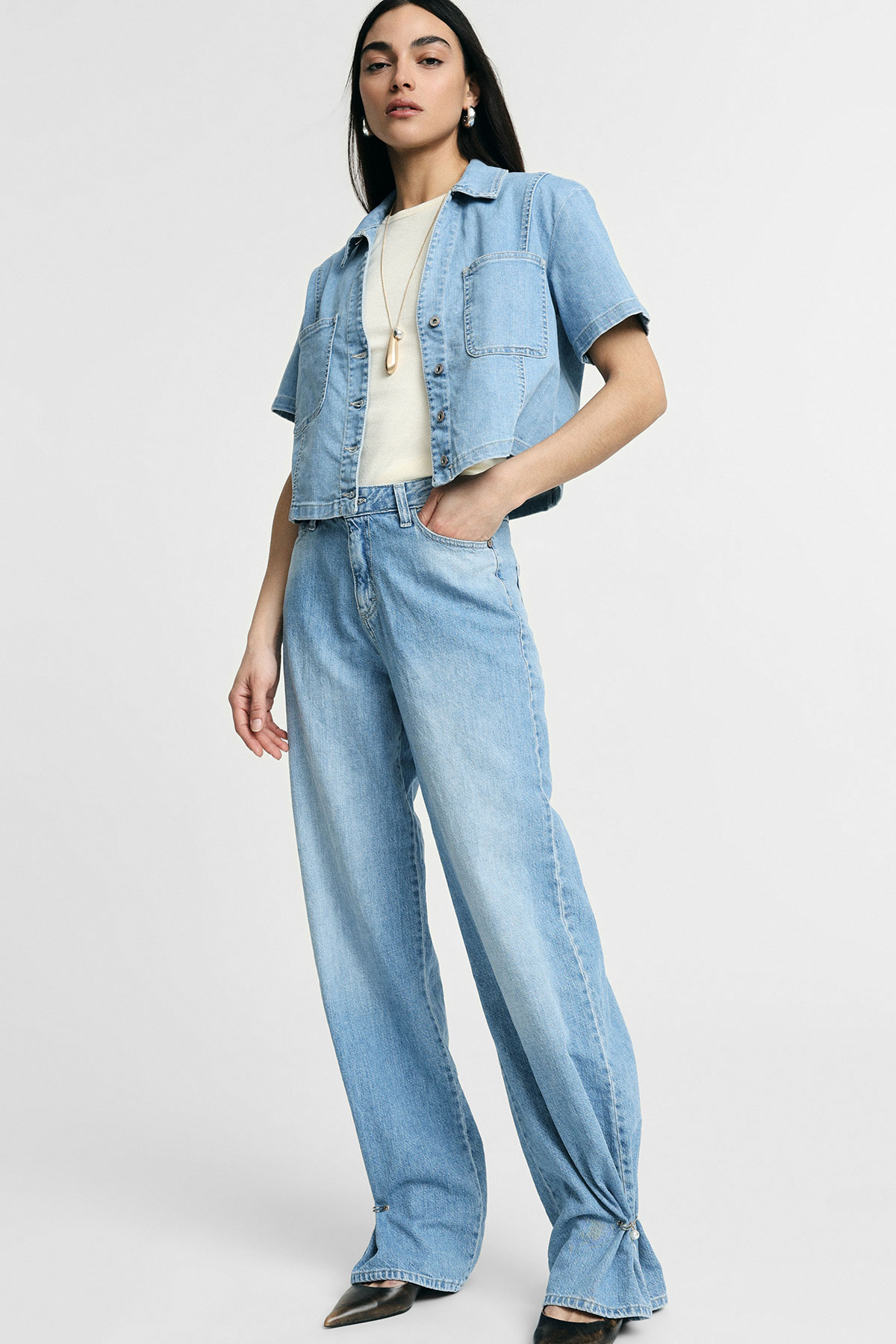 Woman modelling light-wash wide-leg jeans with tied hems, cropped short-sleeve denim shirt, cream top and pendant necklace on neutral background