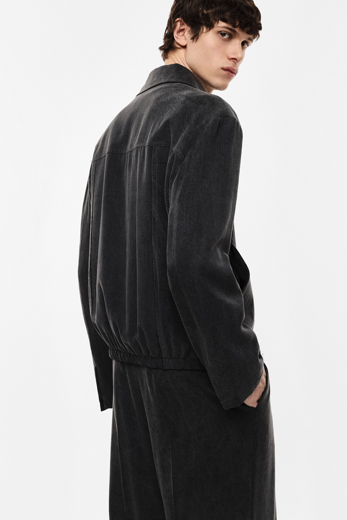 Man in dark charcoal corduroy jacket and matching trousers, three-quarter back pose with hands in pockets on white background