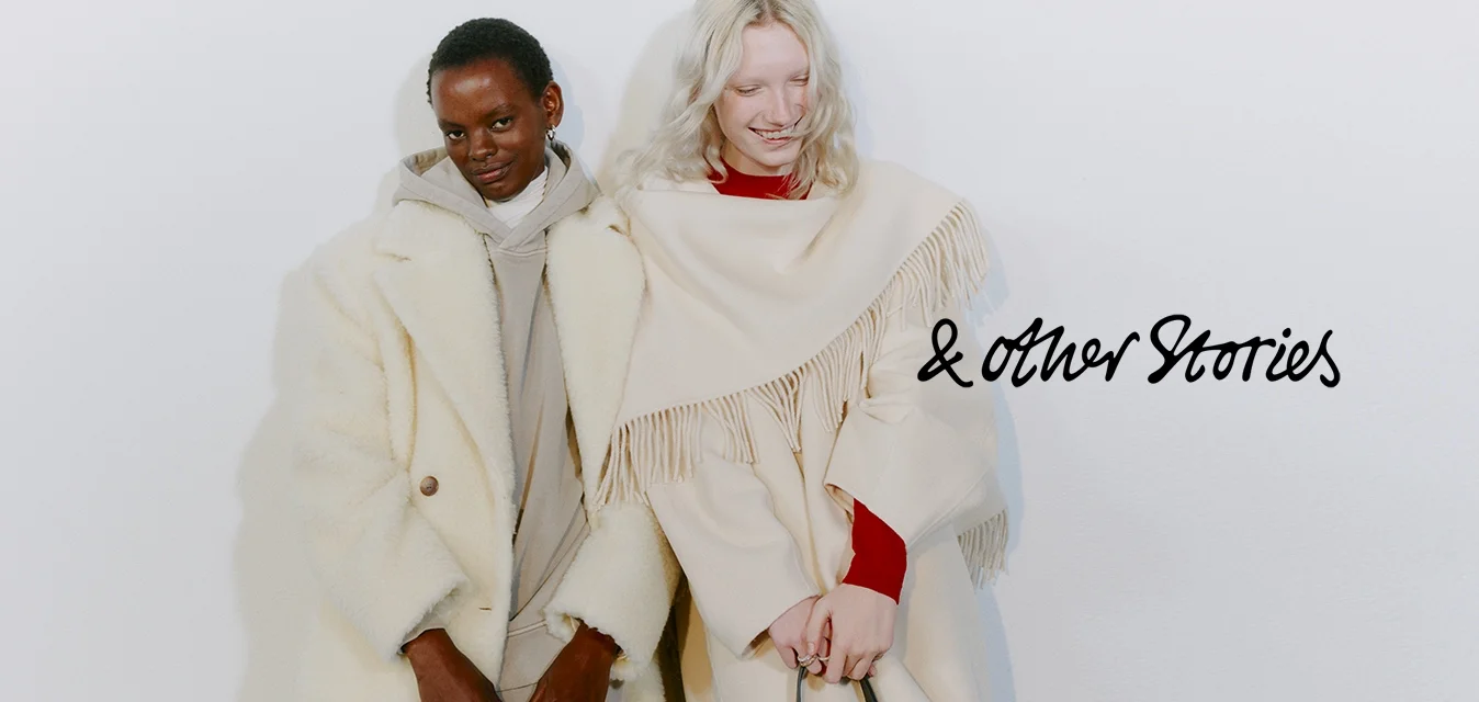 Due persone indossano indumenti esterni di colore chiaro. Una ha un cappotto peloso e testurizzato, mentre l'altra indossa un capo drappeggiato con frange. Entrambi i capi hanno tonalità neutre.