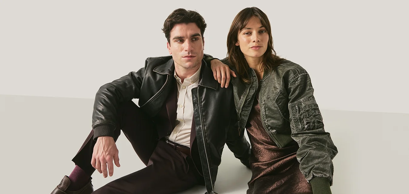 Giovane uomo e donna seduti per terra, uomo con giacca e pantaloni di pelle neri, donna con giacca bomber verde e vestito marrone, sfondo neutro.