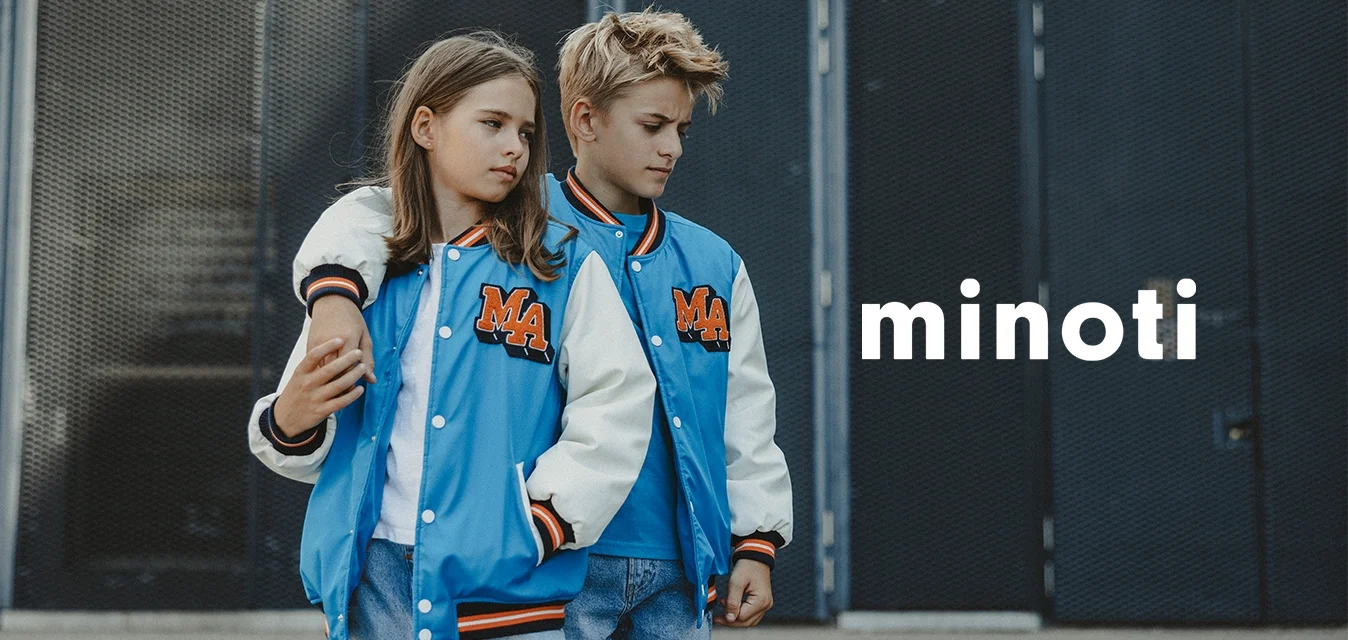 Due bambini indossano giacche varsity blu e bianche abbinate con lettere arancioni. La ragazza ha i capelli lunghi e pantaloni corti; il ragazzo ha i capelli biondi.