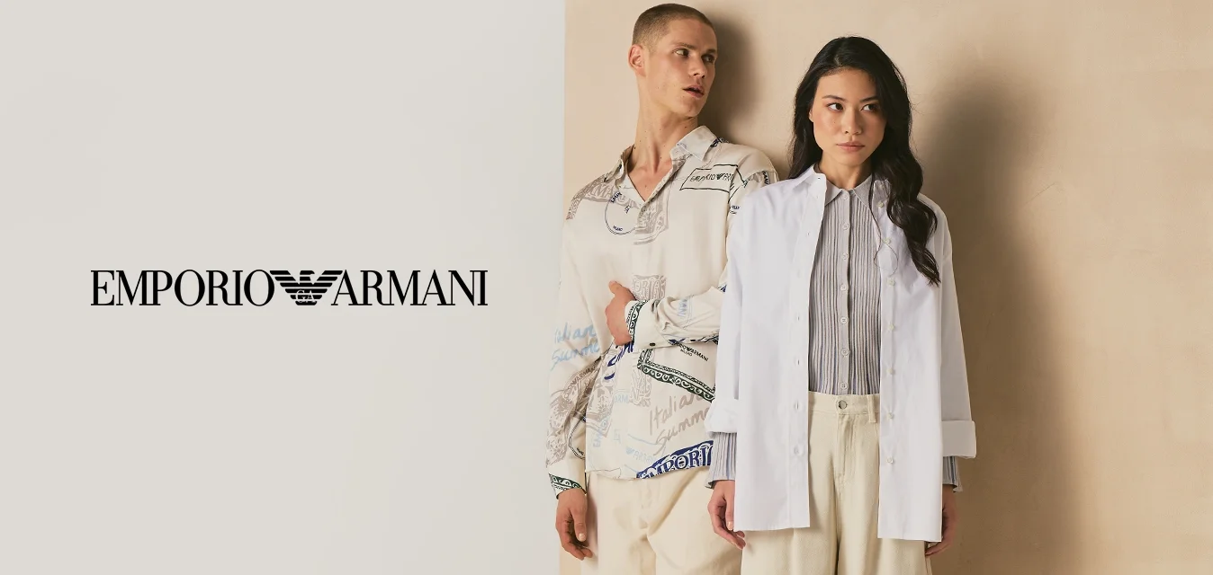 Due persone sono appoggiate a un pannello beige, una indossa una camicia a motivo e pantaloni color crema, l'altra un blazer bianco e pantaloni larghi color crema.