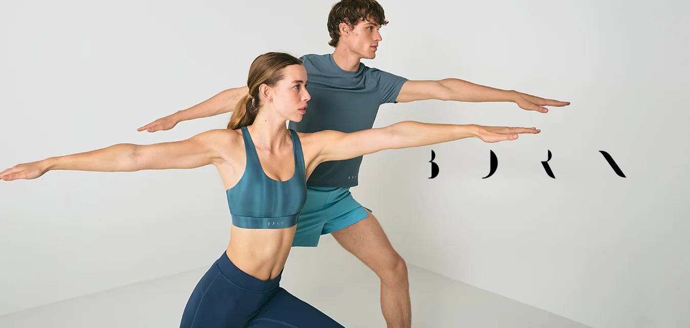 Un uomo e una donna in abbigliamento sportivo eseguono una posa yoga del guerriero, con le braccia tese, in una stanza bianca minimalista.