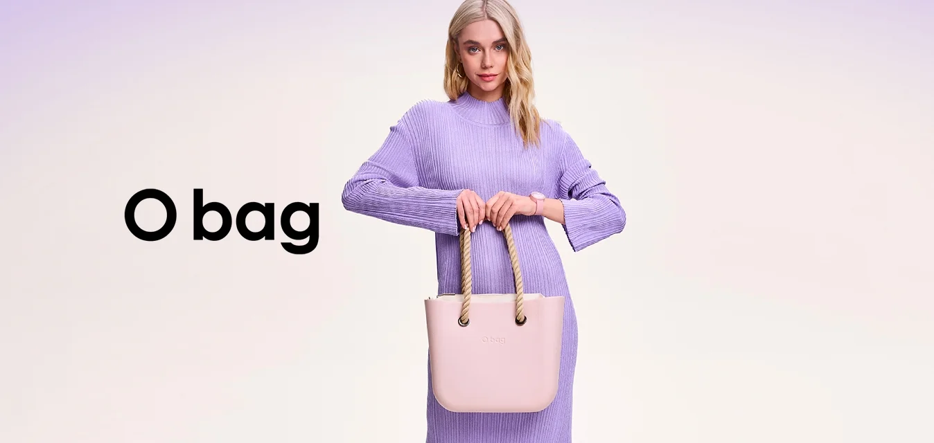 Donna in un lungo abito lavanda che tiene una borsa rosa chiaro con manici in corda, in piedi davanti a uno sfondo chiaro uniforme.