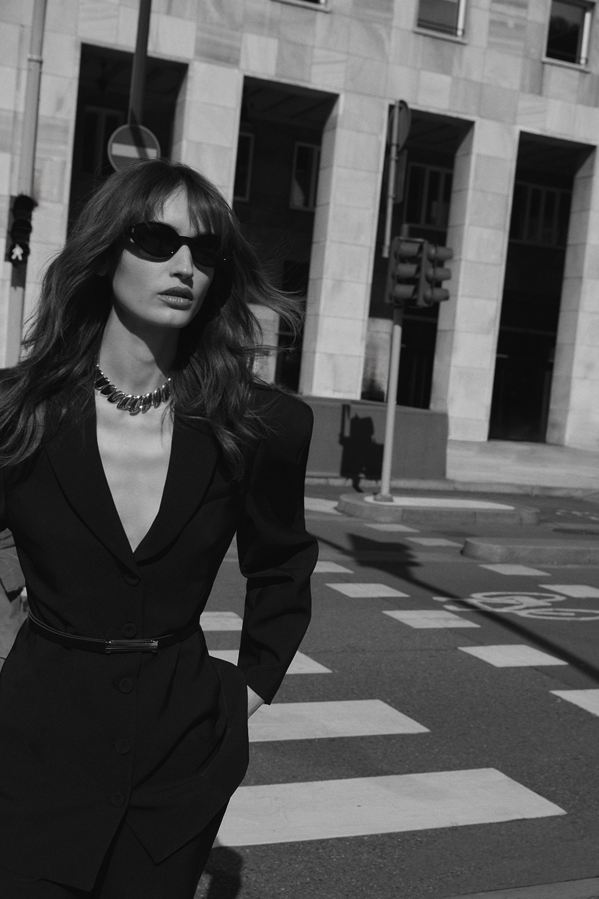 Woman in black blazer and sunglasses walking across a city zebra crossing, statement necklace, monochrome street fashion portrait