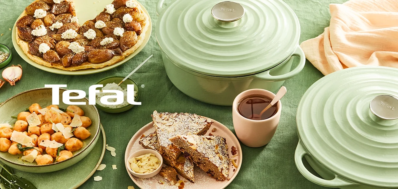 Due contenitori in ceramica verde, una tazza rosa e diversi piatti di cibo disposti su un tessuto verde. Le texture notevoli includono ceramiche lisce e dolci morbidi.