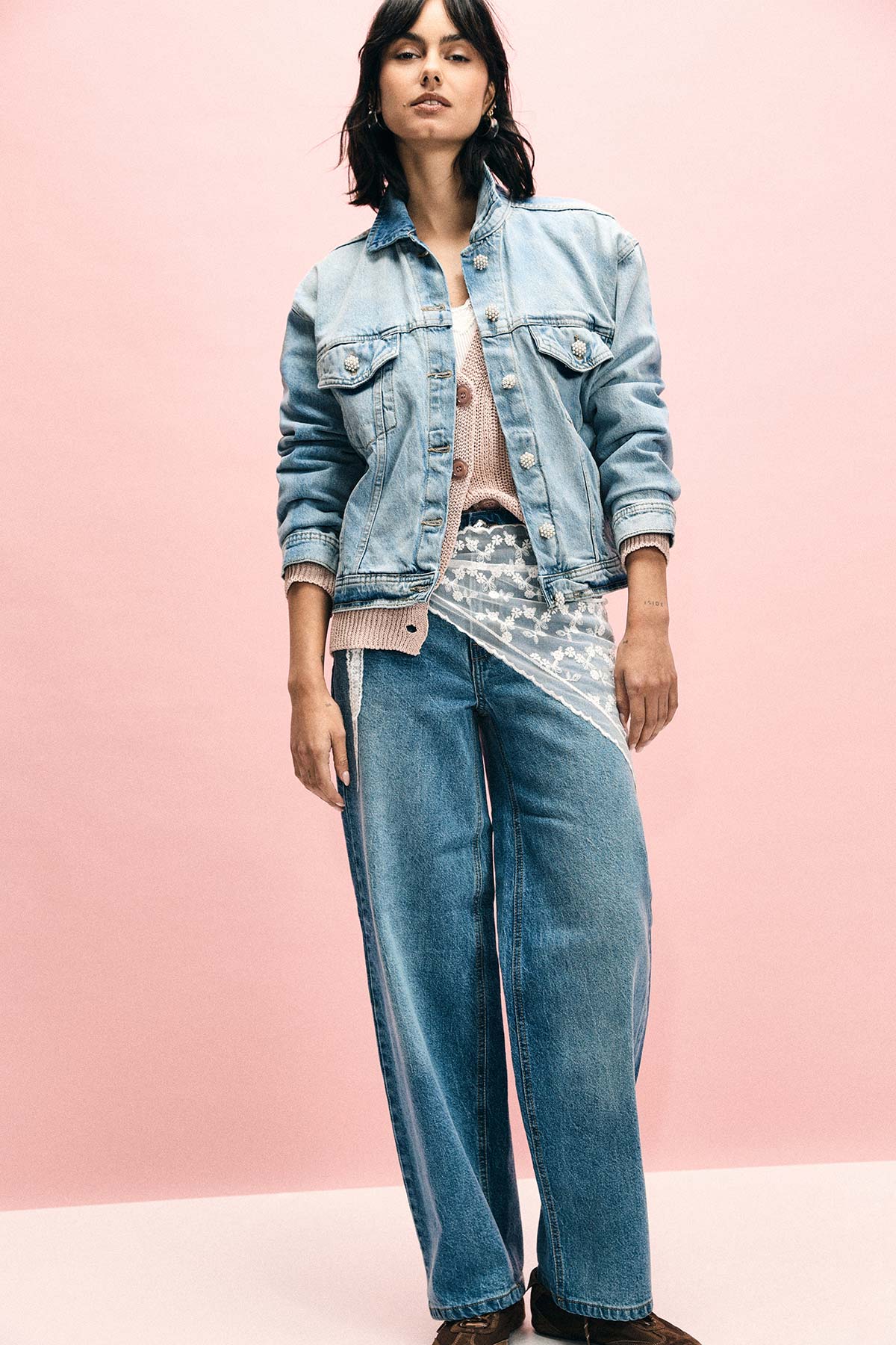 Woman wearing light denim jacket and wide‑leg jeans with layered knit top against pink studio background