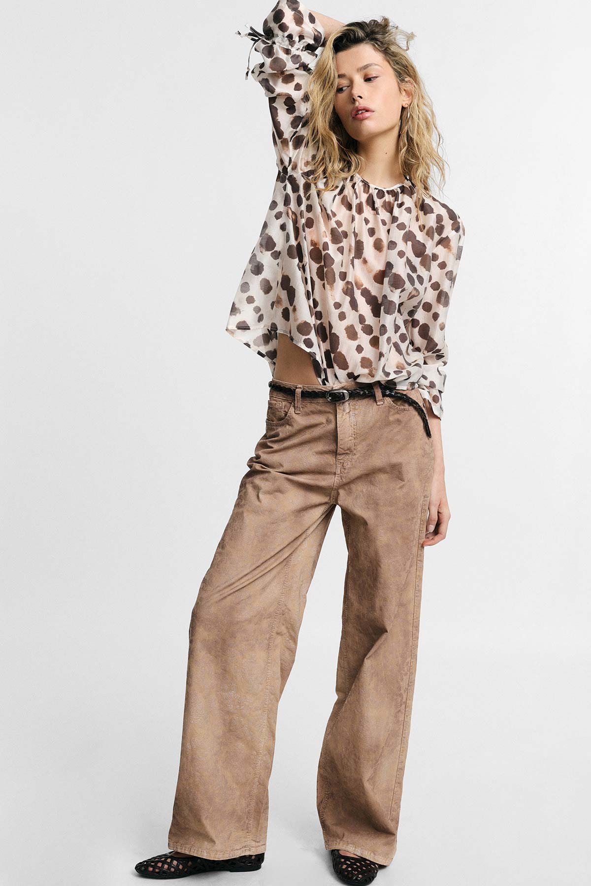 Female model in a sheer white blouse with brown leopard spots and high-waisted wide-leg tan trousers, standing with one arm raised