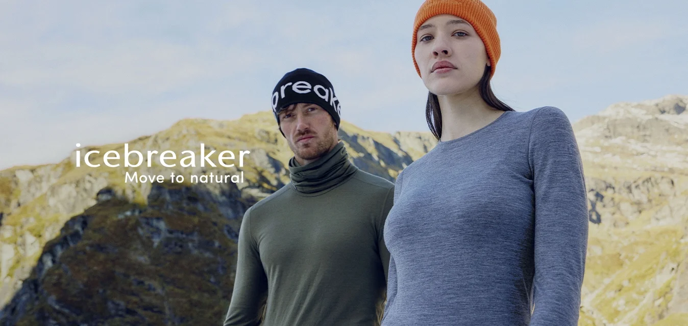 Due adulti con abbigliamento da esterno, indossano strati base con il marchio e berretto, posano davanti a un paesaggio montano sotto un cielo limpido.