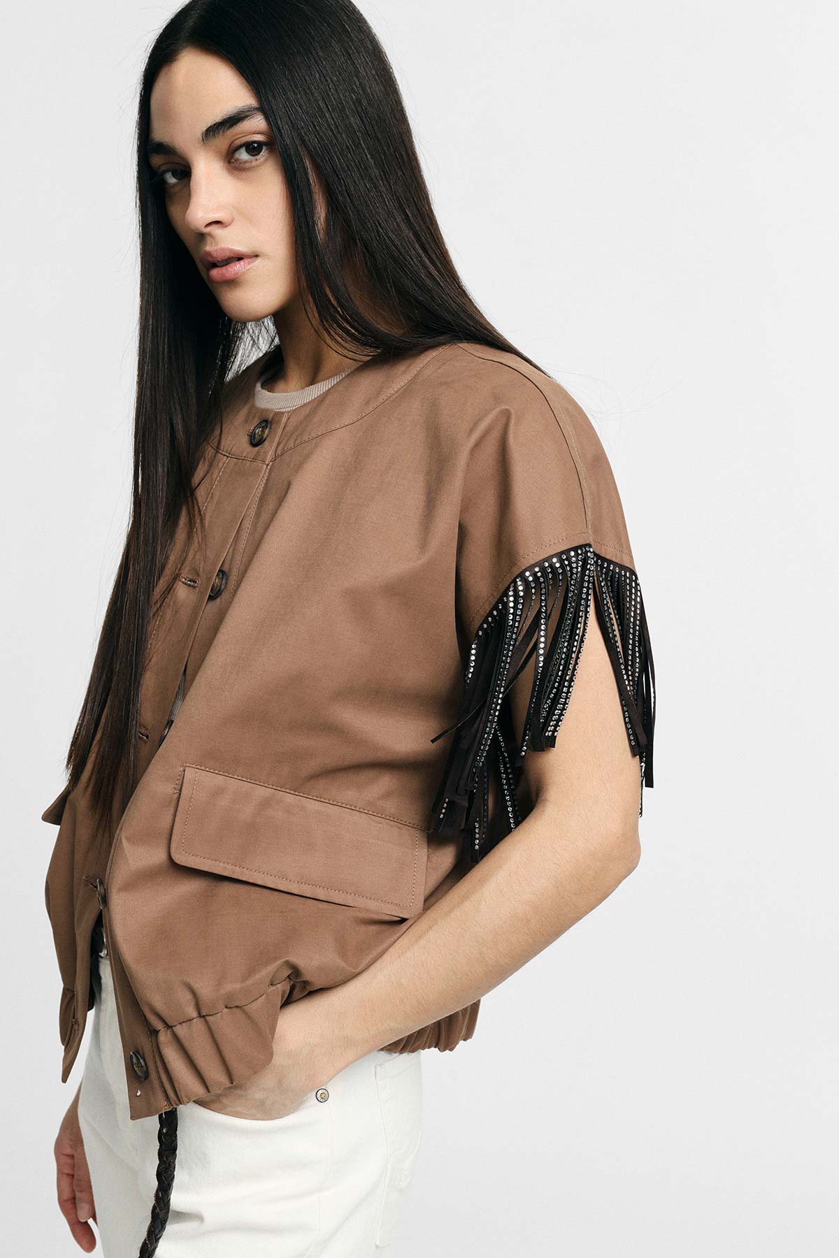 Woman model in brown short-sleeve jacket with black beaded fringe, white trousers and long dark hair against plain light background