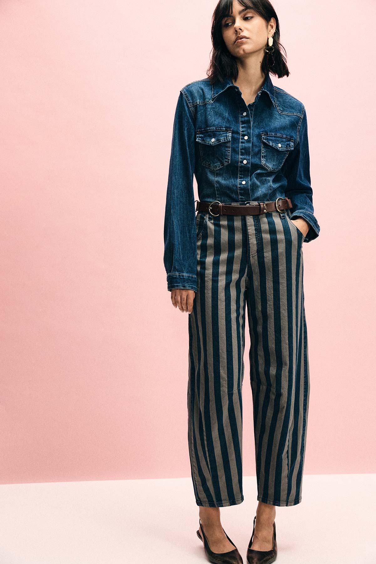 Woman modelling denim shirt and high-waisted navy striped trousers with brown belt against a pale pink background