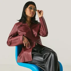 Donna con giacca color bordeaux e pantaloni neri seduta su una sedia moderna blu che si sistema gli occhiali da sole davanti a uno sfondo chiaro uniforme.