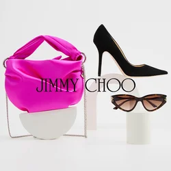 Borsa in satin rosa con manico annodato, décolleté nere a punta e occhiali da sole in acetato tartarugato esposti su stand bianchi astratti.