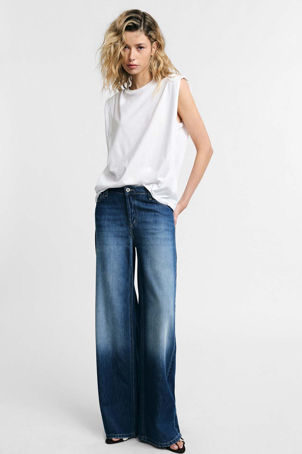 Woman wearing a white sleeveless top and high-waisted wide-leg blue jeans, hands in pockets against a plain studio background