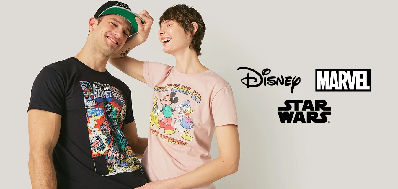 Due giovani adulti sorridenti, uno indossa una maglietta nera a tema fumetti e un cappellino, l'altro si sta sistemando il cappellino e indossa una maglietta rosa con un personaggio dei cartoni animati.