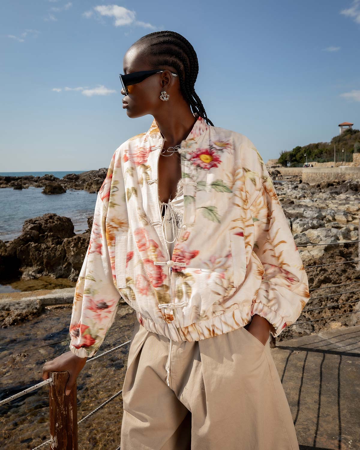 Model in floral bomber jacket, beige wide-leg trousers and sunglasses posing on rocky seafront with blue sky