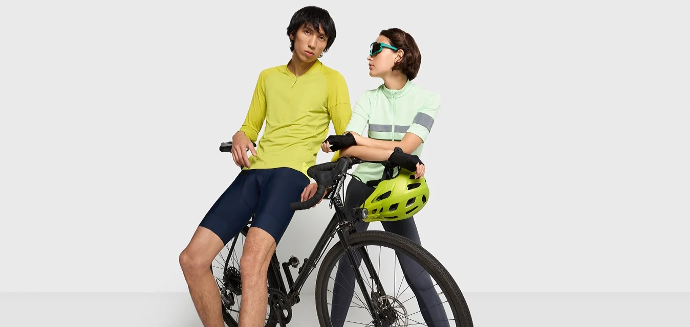 Attrezzatura per il ciclismo mostrata: una persona con una maglietta gialla e pantaloni corti blu scuri, un'altra con una maglietta a righe verde chiaro e pantaloni grigi, con un casco verde su una bici nera.