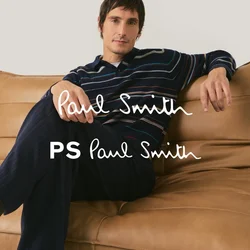 Uomo con maglietta a righe a maniche lunghe e pantaloni scuri seduto a gambe incrociate su un divano in pelle color cuoio con sfondo semplice.