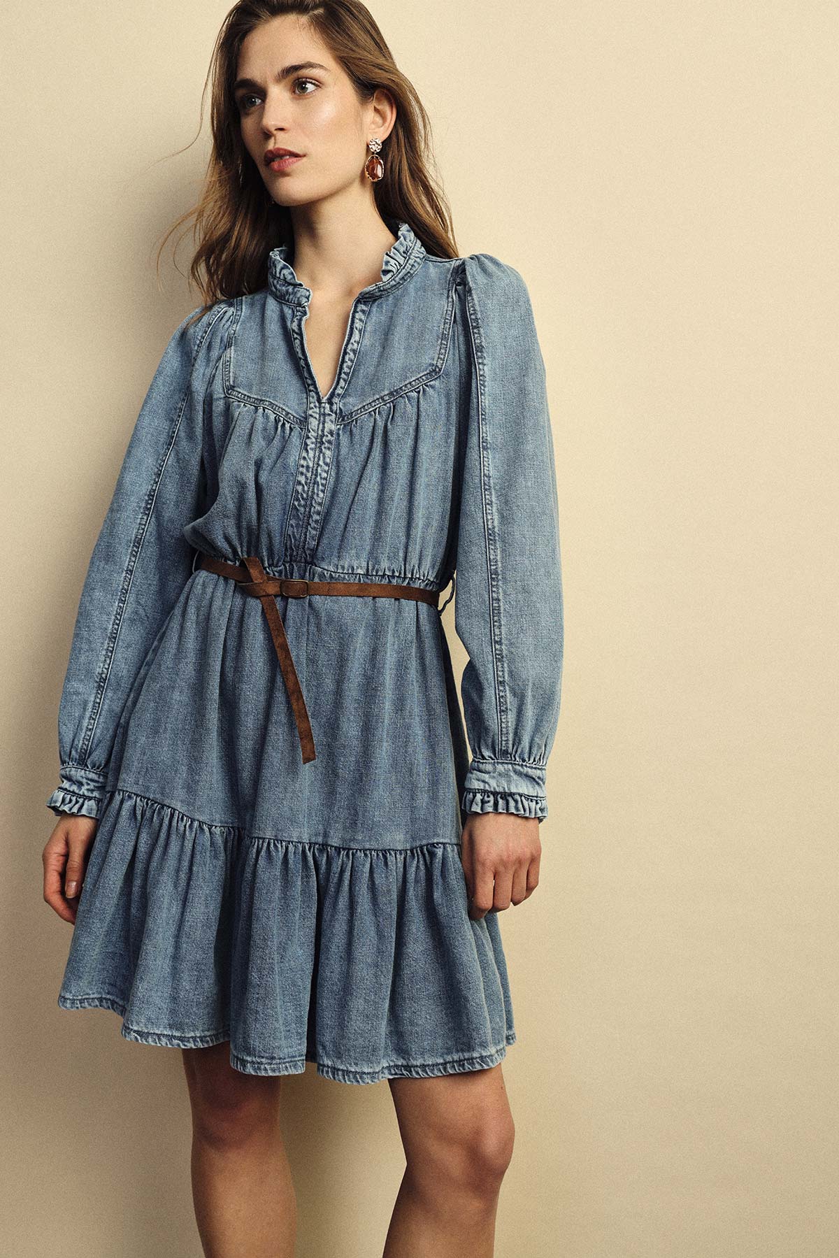 Woman wearing a blue denim long-sleeve ruffled-collar dress with a tiered skirt and thin brown belt, standing against a beige background