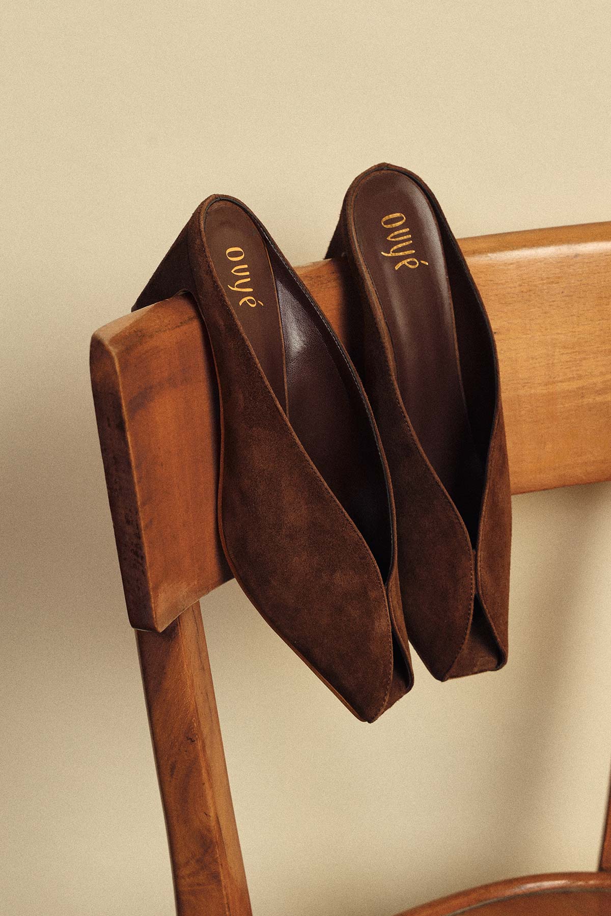 Pair of brown suede pointed-toe flats draped over a wooden chair, minimalist product shot