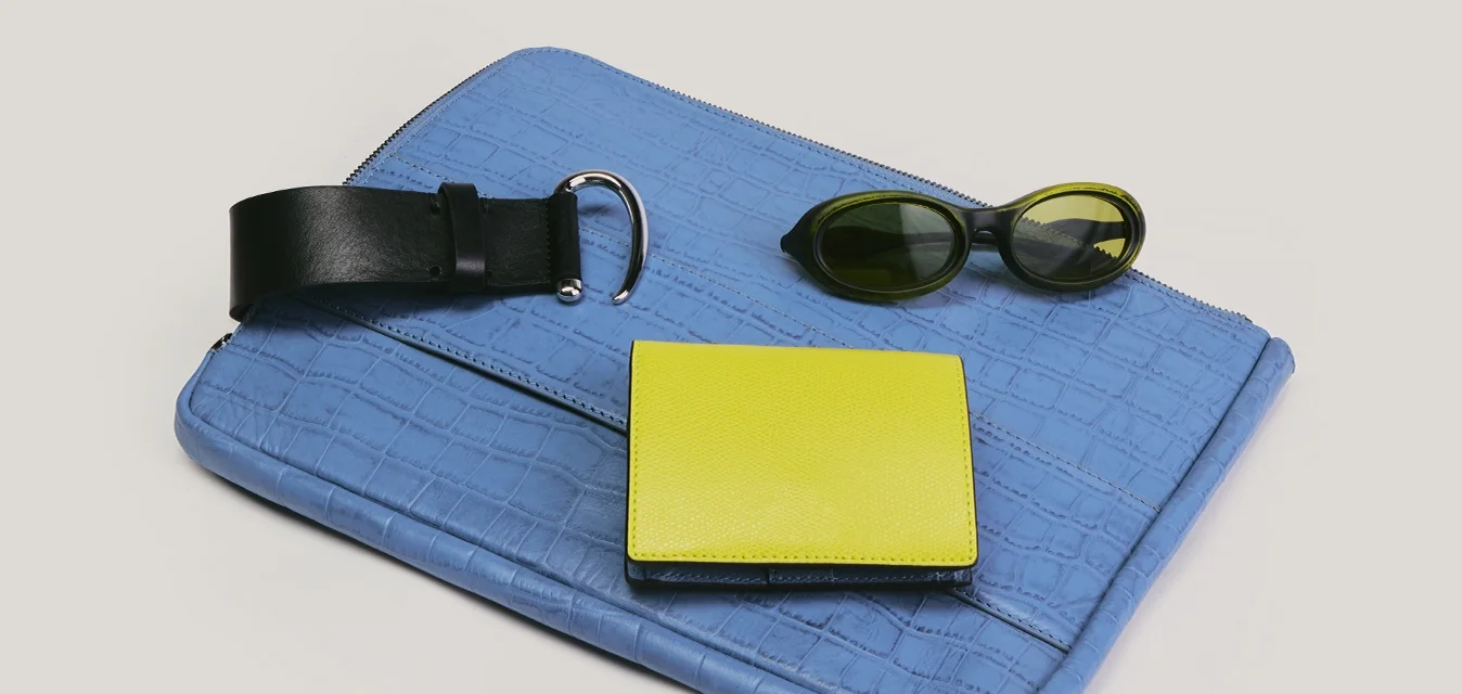 Clutch blu testurizzato con cintura in pelle nera, portafoglio giallo e occhiali da sole ovali scuri disposti su uno sfondo chiaro.