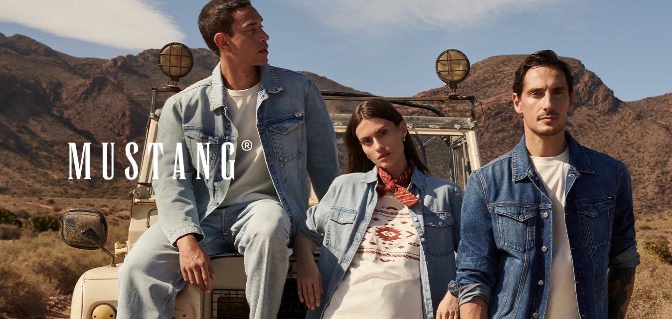 Tre persone in abiti di jeans posano accanto a un veicolo fuoristrada su un sentiero sterrato con un paesaggio desertico montuoso sullo sfondo.