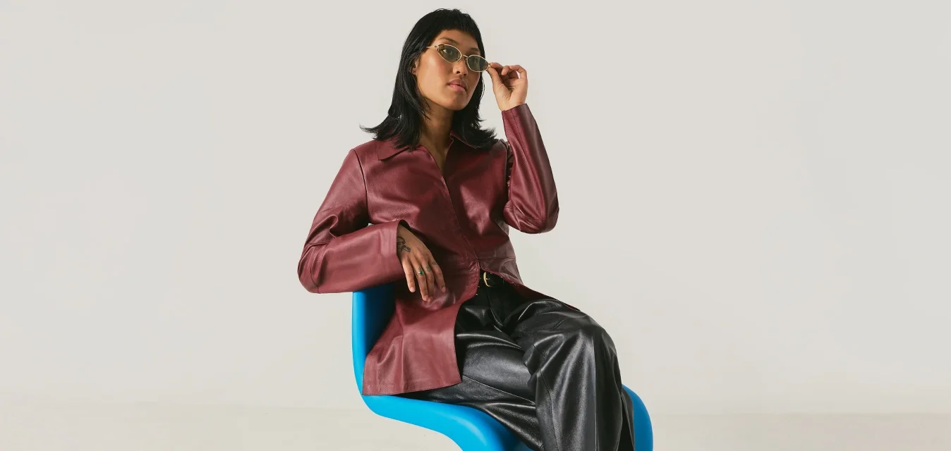 Donna con giacca color bordeaux e pantaloni neri seduta su una sedia moderna blu che si sistema gli occhiali da sole davanti a uno sfondo chiaro uniforme.
