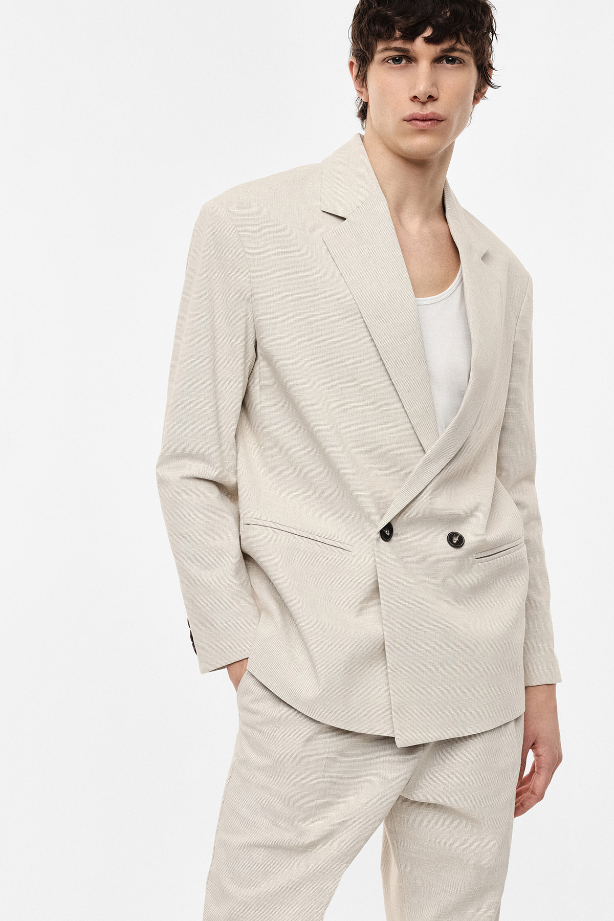 Male model in beige double-breasted suit and white vest top, hands in pocket, standing against plain white background