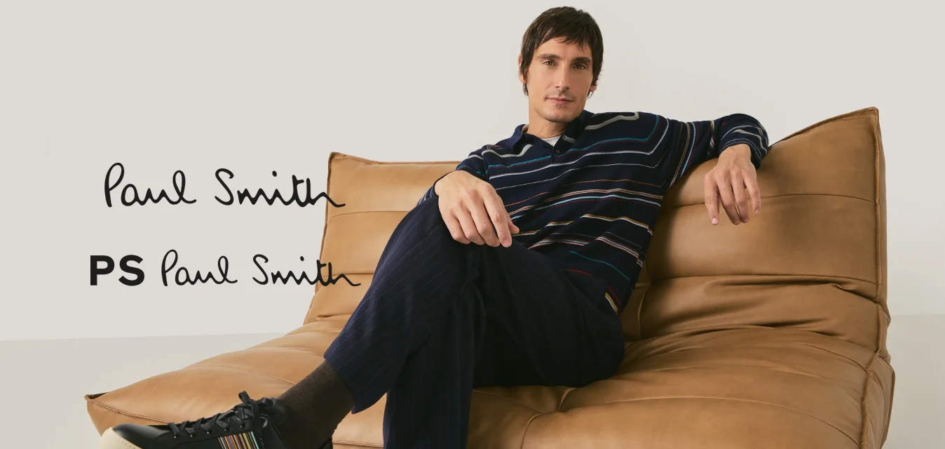 Uomo con maglietta a righe a maniche lunghe e pantaloni scuri seduto a gambe incrociate su un divano in pelle color cuoio con sfondo semplice.