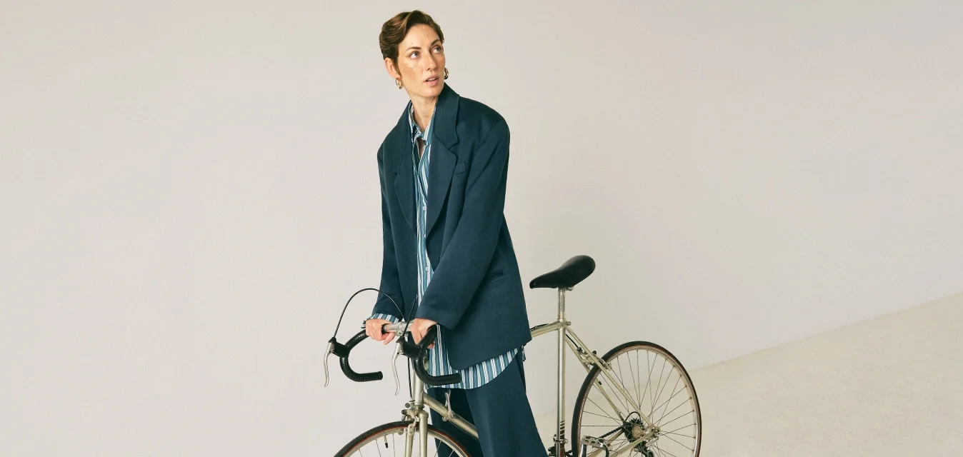 Donna in un abito blu scuro in piedi mentre tiene una bicicletta da strada vintage con manubri a caduta su uno sfondo beige semplice.