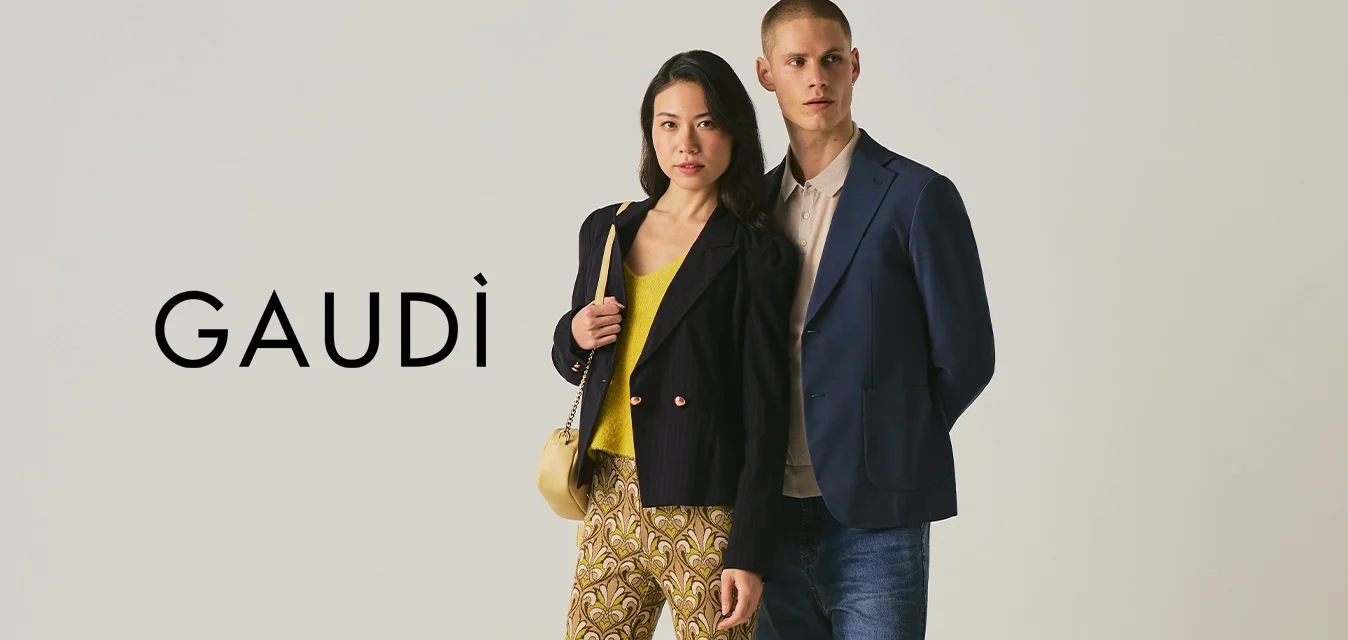 Donna con pantaloni a fantasia e blazer nero accanto a uomo con blazer blu e jeans su sfondo neutro.