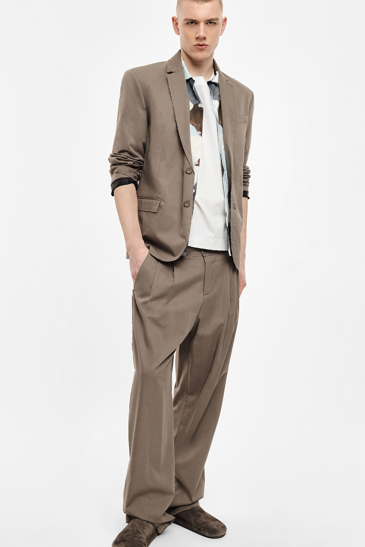 Model wearing taupe relaxed-fit suit with rolled sleeves, patterned shirt over white tee, hands in pockets, brown suede shoes