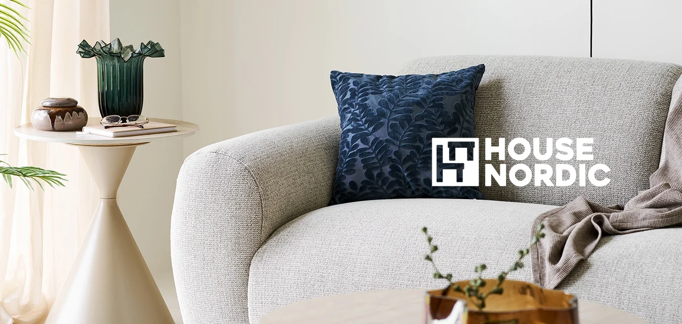 Divano curvo grigio chiaro con cuscino blu scuro a motivo foglia e coperta beige, tavolino laterale e da caffè beige con oggetti decorativi in un soggiorno luminoso.