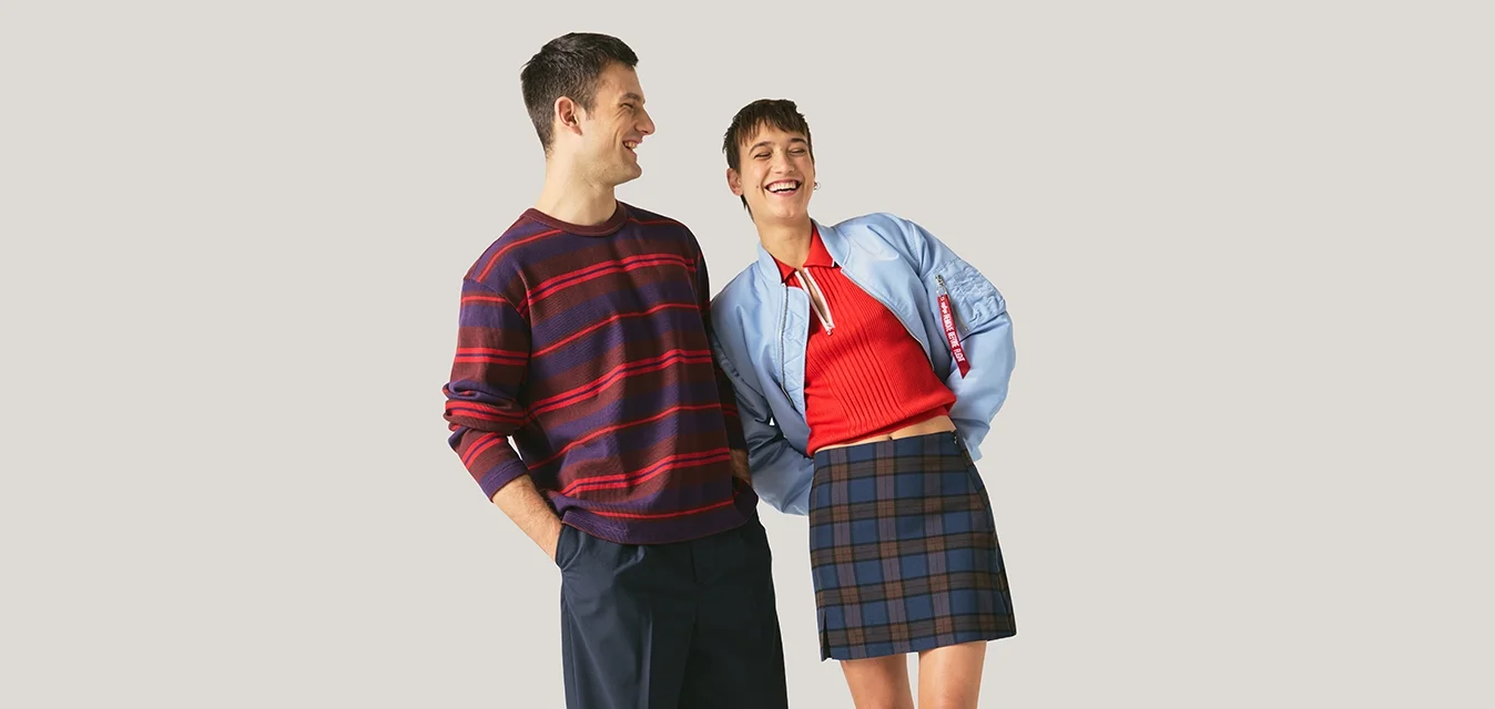 Uomo con maglione a righe e pantaloni scuri sorride accanto a una donna con top rosso, giacca blu, gonna a quadri e stivali neri su sfondo neutro.