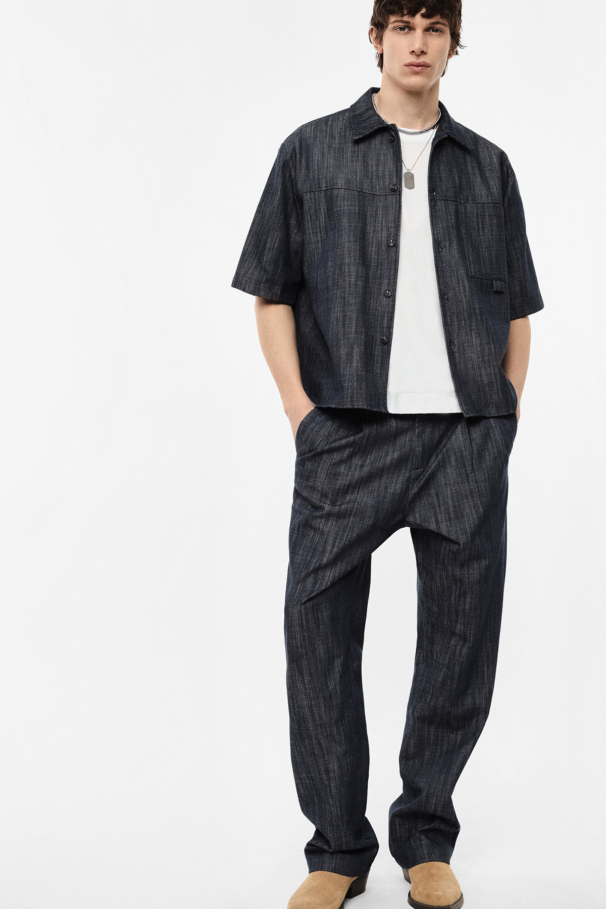 Male model wearing dark denim short-sleeve overshirt with matching relaxed trousers, white T-shirt and tan suede shoes
