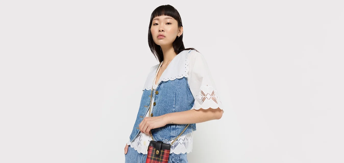 Salopette di denim con un top in pizzo bianco, accompagnata da una piccola borsa a tracolla a fantasia scozzese rossa. L'outfit combina diverse trame di tessuto blu e motivi contrastanti.