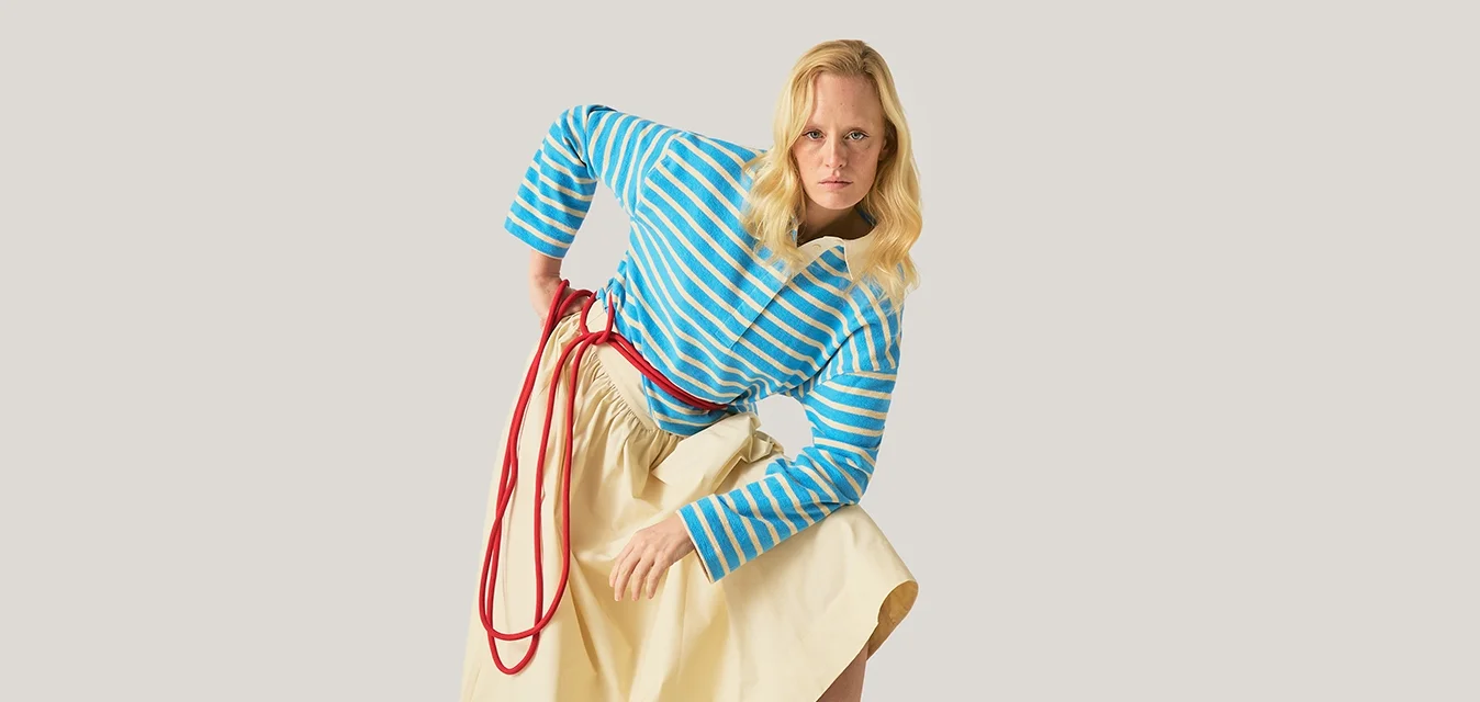Donna bionda con un maglione a righe blu e una gonna beige si sporge in avanti con un piede su una cassa di legno, sullo sfondo neutro.
