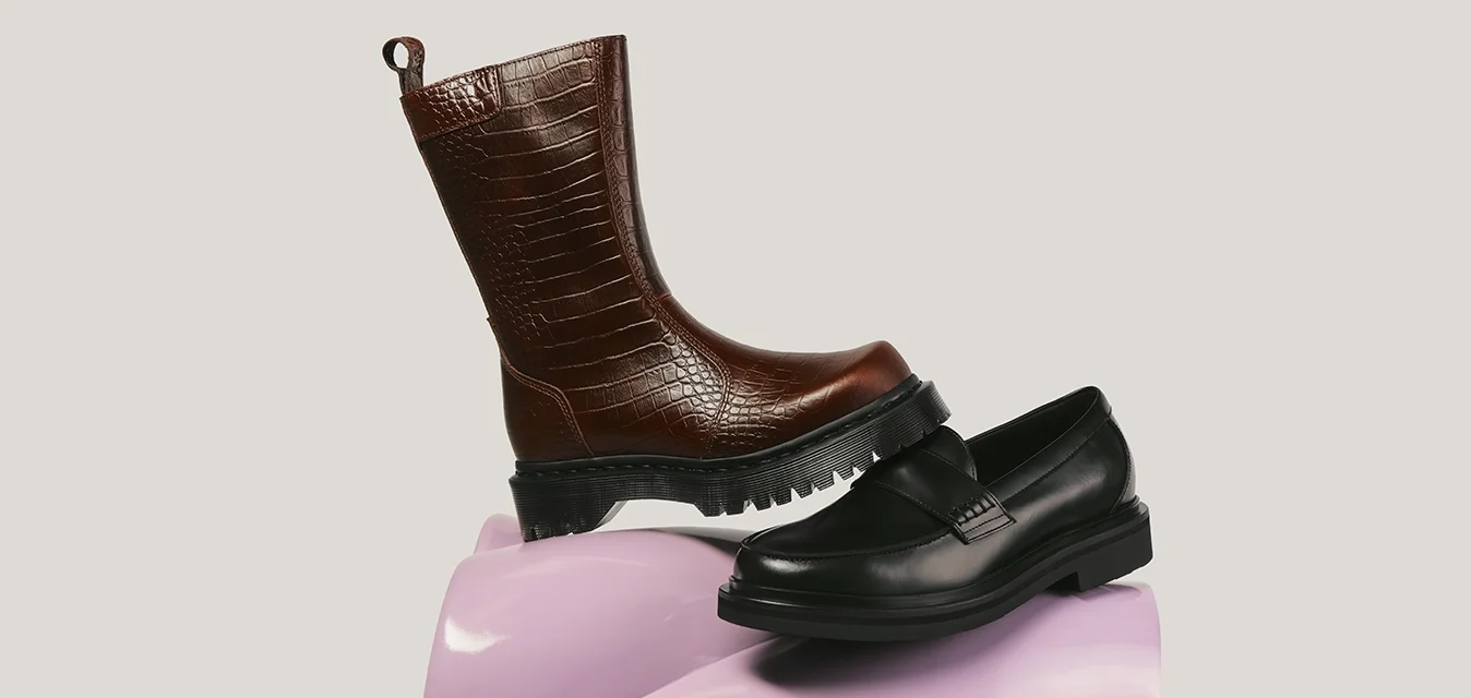 Stivale marrone in pelle con motivo coccodrillo e mocassino nero in pelle esposti su un piedistallo rosa lucido e tortuoso.
