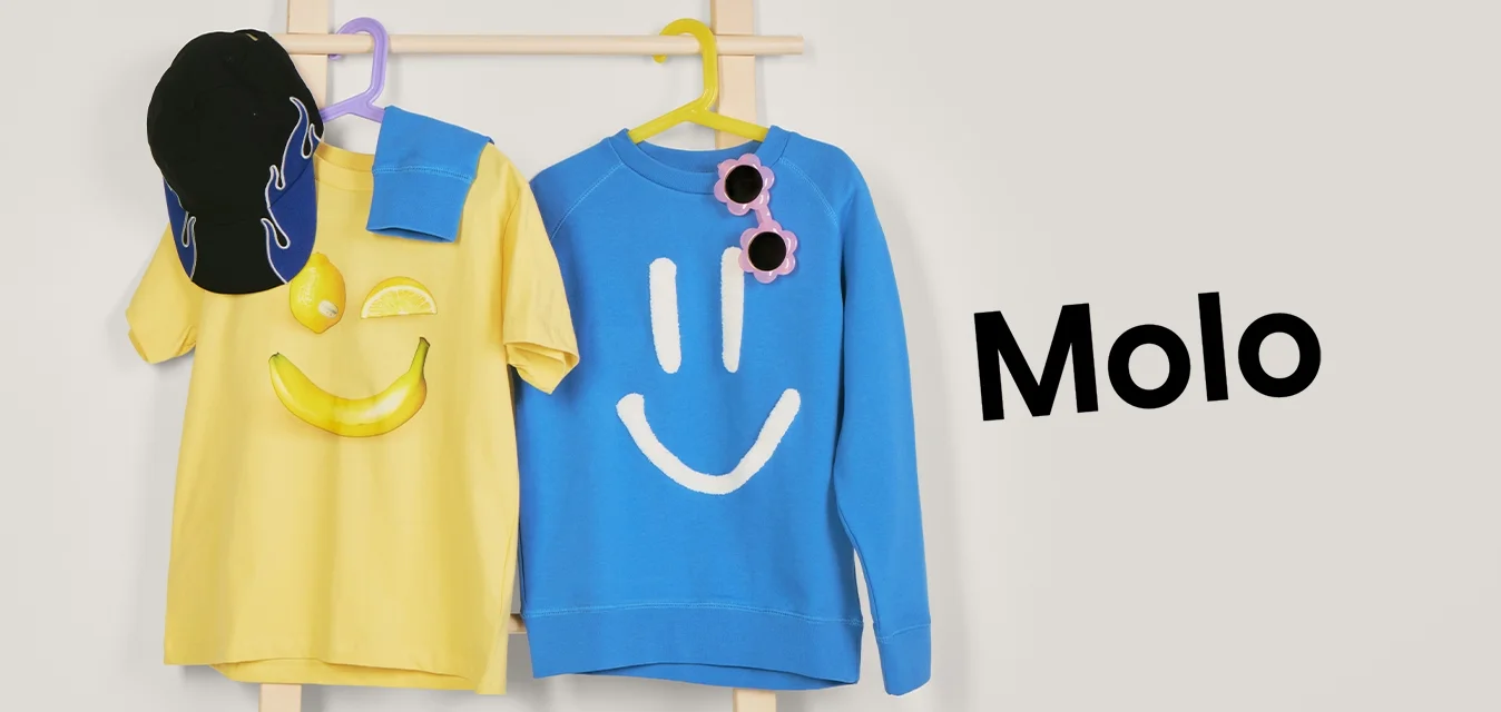 Maglietta gialla con faccina sorridente e cappellino nero, e felpa blu con faccina sorridente bianca e occhiali da sole rosa appesi a un attaccapanni di legno.