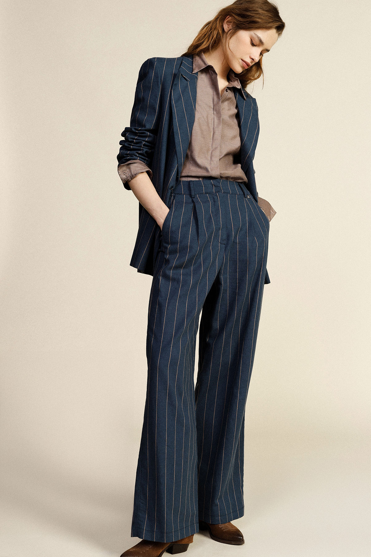 Woman wearing navy pinstripe blazer and wide-leg trousers with brown blouse and shoes, hands in pockets, looking down