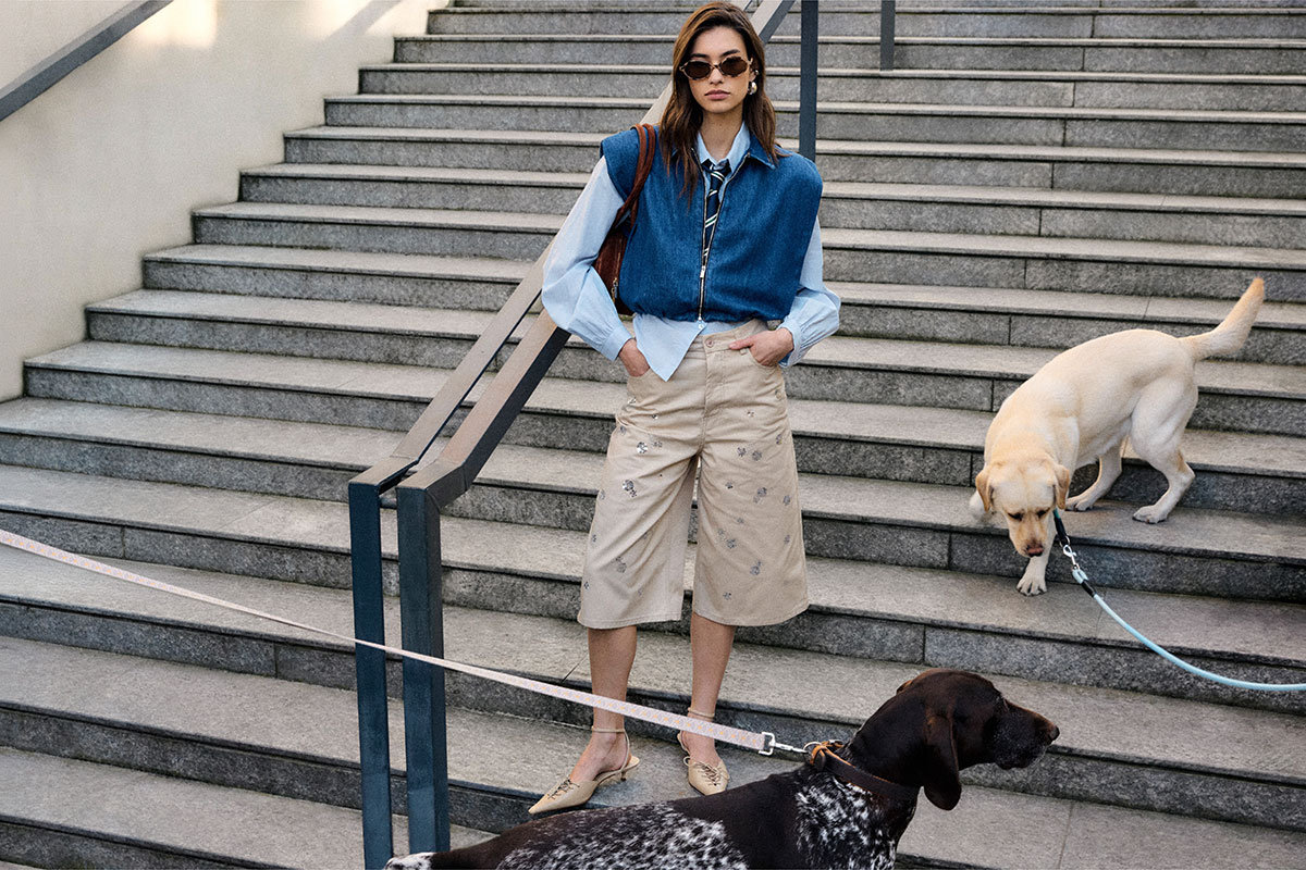 Woman in sunglasses and denim vest standing on outdoor steps with two leashed dogs, wearing beige culottes